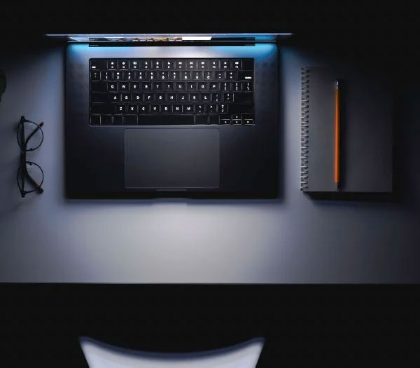 Backlit laptop keyboard on a dark desk beside glasses and a notebook